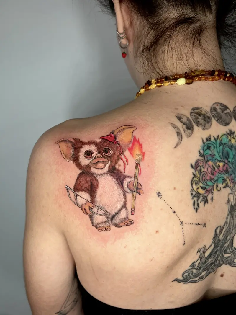 Tatuaje a color de criatura tipo gremlin adorable con lazo, sosteniendo una antorcha y un cuchillo.