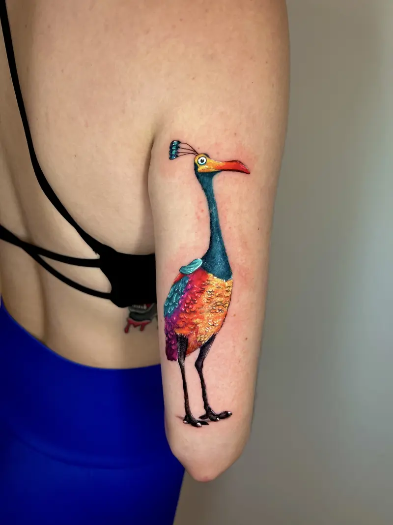 Tatuaje a color de un ave estilizada con plumaje vibrante y diseño ilustrativo en la parte posterior del brazo.